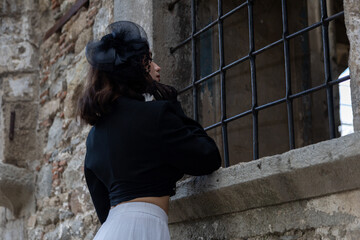 Young woman with black hair, dressed in black Gothic style, in a dilapidated church with barred windows.