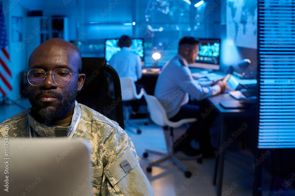 Aufkleber serious young man in camouflage uniform sitting in front of desktop computer against coworkers in mi