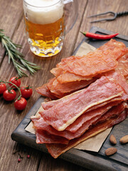 Smoked pork and chicken meat chips on a wooden tray with vegetables and a glass of light beer on the table