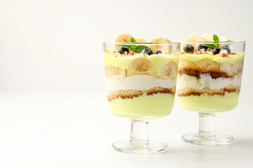 Tasty trifle dessert. Banana, blueberries, sponge cake and whipped cream in glasses on white wooden table, space for text