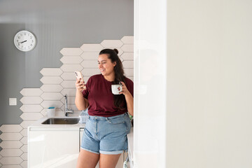 Curvy woman enjoys morning coffee while using smartphone in kitchen