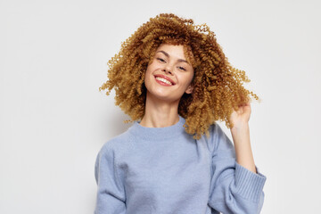 Smiling young woman with curly hair in a light blue sweater against a neutral background,...