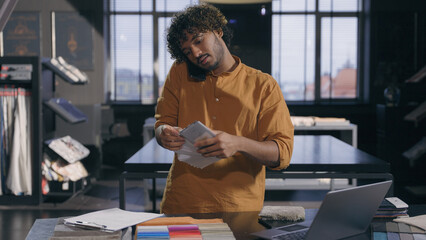 Arabian Indian male business man working busy interior designer talking with client online discuss on mobile phone fabric tissue samples write in notepad textile order in workshop modern design studio