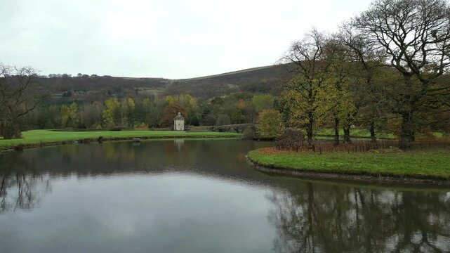 Low drone flying over tranquil small lake towards an old stone bridge in Holmfirth town, England, UK