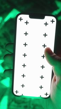 Vertical video. Close-Up of a Hand Swiping Downward on a Smartphone with a Blank White Screen Mockup and Tracking Markers, Positioned Over a Green-Glowing Background of US Hundred-Dollar Bills, Repres