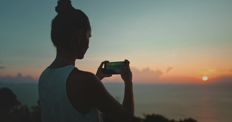 Capturing the sunset at the beach with a smartphone, a woman enjoys the colorful sky reflecting on the ocean, creating lasting memories of her travels