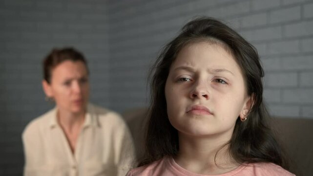 Woman scolding bored daughter. A view of bored little girl sitting by the screaming mother on the sofa.