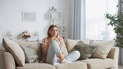 Frightened woman is watching a scary movie on tv while sitting on a sofa in her living room, hugging her knees and experiencing intense fear and anxiety