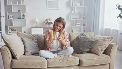 Young woman is sitting on comfortable sofa at home, raising her fist in excitement while looking at her smartphone, celebrating online win, successful purchase or good news