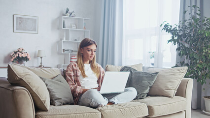 Comfortable young woman sitting on a sofa with crossed legs using a laptop in her living room, working from home in a bright and cozy environment