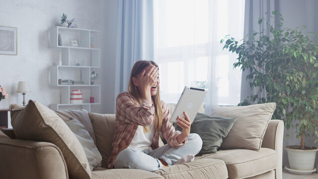 Young woman sitting on a comfortable sofa at home, holding a tablet and reacting with excitement and surprise to good news, covering her face with one hand