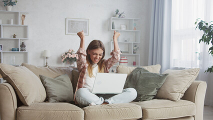 Fototapeta premium Young woman sitting comfortably on a sofa, laptop resting on her lap, raising her arms in joyful celebration of success while working from home in a cozy living room setting