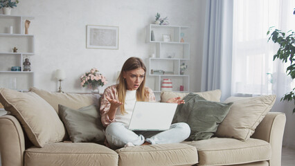 Stressed young woman sitting on a cozy sofa, raising hands in shock while staring at her laptop screen, reacting to unexpected online news