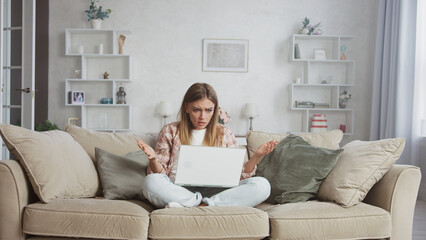 Young woman sitting with crossed legs on a beige sofa in her living room, looking confused and...
