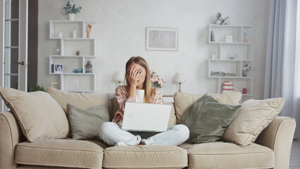 Young woman sitting on a sofa with crossed legs working with a laptop and covering her face with...