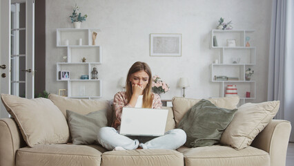 Young woman sitting on the sofa with crossed legs is covering her mouth with her hands and crying, reading bad news on a laptop, having a mental breakdown