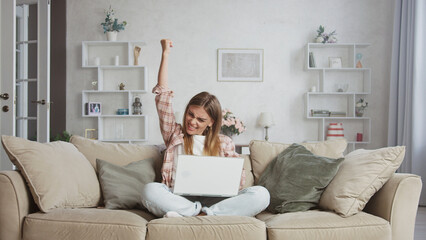 Young woman raising her arm in excitement while using a laptop on the sofa, celebrating an online...