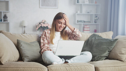 Young happy woman working from home while sitting on a comfortable sofa, using her laptop and feeling surprised and excited about her recent online win