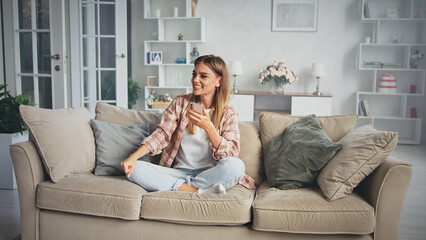 Young woman sitting comfortably on a cozy sofa in a modern living room, using her smartphone while smiling and glancing away, enjoying a moment of relaxation and connection