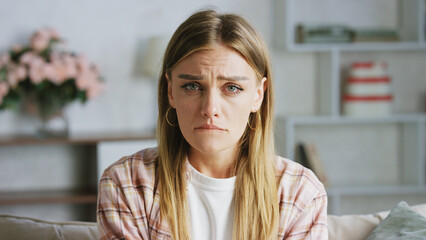 Portrait of a young woman sitting on a sofa in her living room, expressing deep sadness and...