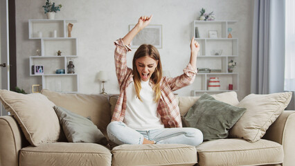 Overjoyed young woman sitting with crossed legs on comfortable couch, raising arms and shouting...