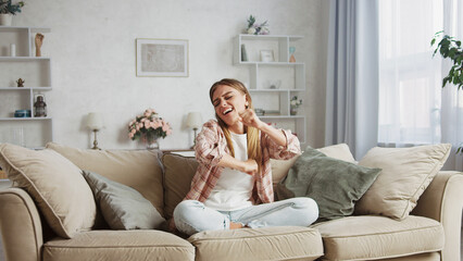 Happy young woman sitting cross legged on a comfortable sofa, singing and dancing joyfully while enjoying music in a cozy living room filled with natural light