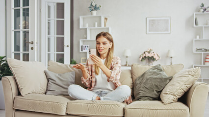 Young woman making a video call using her smartphone while sitting comfortably on a sofa in her modern, bright living room, engaging in a conversation