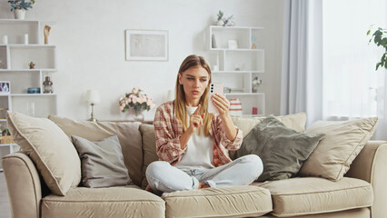 Young woman sitting with crossed legs on comfortable sofa in cozy living room, making video call using smartphone, pointing finger at screen while talking to someone online