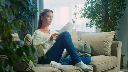 Fototapeta premium Young woman wearing jeans and white sweater is sitting comfortably on sofa in living room, reading a document with focused expression, surrounded by houseplants, enjoying quiet moment at home