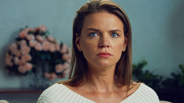 Portrait of a serious woman with blue eyes showing determination and confidence while sitting on a sofa, with a blurred bouquet of flowers in the background, in a softly lit environment
