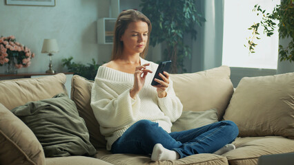 Obraz premium Young woman is using mobile phone while sitting comfortably on sofa in living room, browsing internet or social media, enjoying leisure time at home