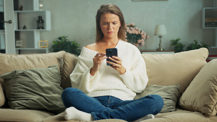 Fototapeta premium Young woman is reading shocking news on smartphone sitting on sofa at home, she is feeling frustrated and confused, having problems and troubles
