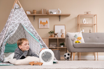 Cute little boy lying in play tent near electric fan heater at home