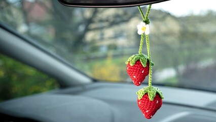 In a car interior, two crochet strawberries, complete with green leaves and tiny white flowers,...
