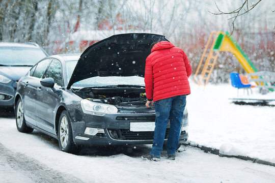 Сar breakdown, Man in red jacket checking car engine under an open hood during snowfall. Vehicle troubleshooting, man inspects car engine under cold weather conditions. Fluid levels control in winter.
