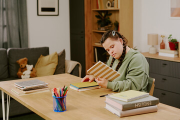Modern Caucasian girl with Down syndrome holding notebook sitting at table in living room at home,...