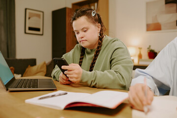 Medium shot of Caucasian girl with Down syndrome sitting at desk in living room, checking message on smartphone, hand of unrecognizable woman writing in notebook