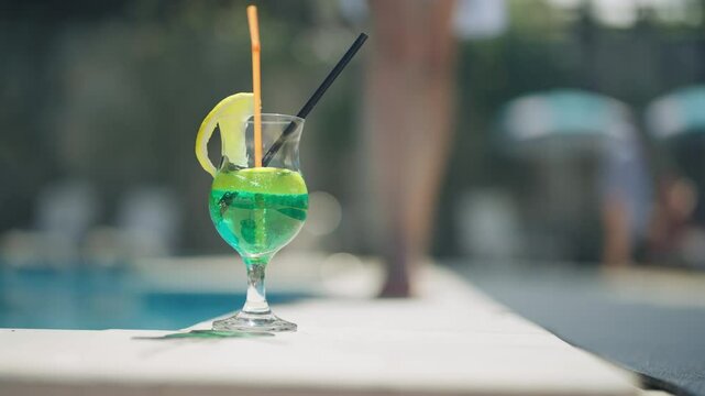 A vibrant green mojito cocktail with a lemon slice and colorful straw, set against a blurry poolside background with walking sexy woman