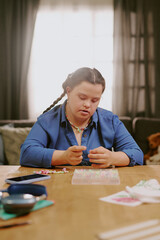 Vertical shot of creative young woman with Down syndrome sitting at table in living room beading bracelet, copy space