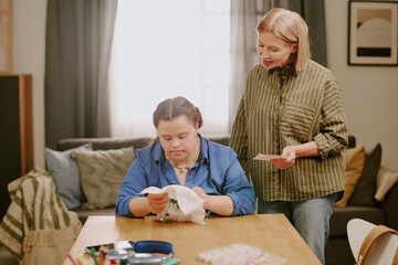 Mature Caucasian woman standing at desk in living room, watching her daughter with Down syndrome...