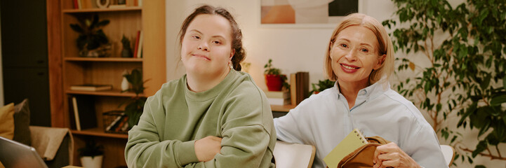 Portrait of young student with Down syndrome and her mature mother sitting at table and smiling at...