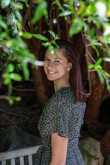 A smiling woman stands in a serene outdoor setting, framed by soft natural light and lush greenery. Her floral dress and gentle expression exude warmth, blending beautifully with the natural backdrop
