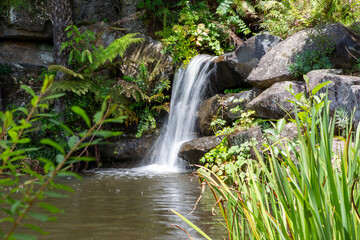 A serene waterfall cascades over mossy rocks into a calm pond, surrounded by lush greenery and vibrant plants. The natural beauty and tranquil ambiance create a picturesque, refreshing scene