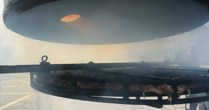 Meat smoking on a large outdoor circular grill, enveloped in a dense, aromatic smoke at a food festival.