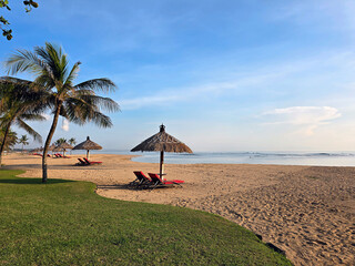 Beach lounge bench with straw umbrella on tropical beach with blue sky background, concept of summer vacation in Bali with negative space