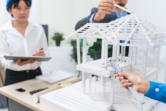 businessmen and a woman wearing hats collaborate at a desk in an office,designing a model house structure,discussing roof designs, accepting concepts,and focusing on teamwork and building innovation