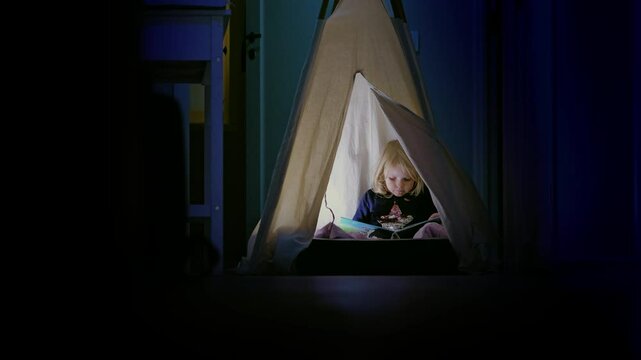 Cute little girl reading alone at night in a tent at home