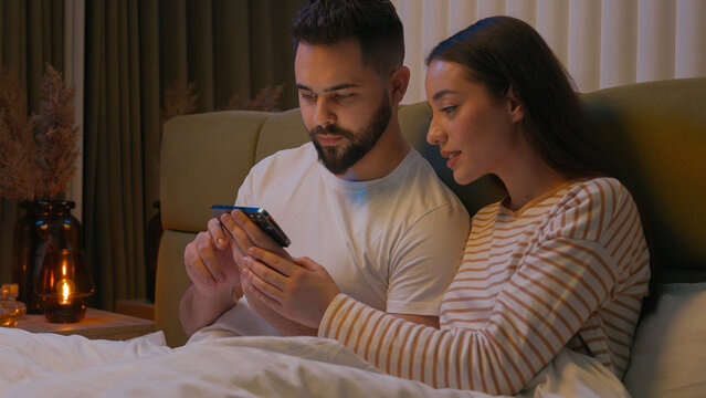 Caucasian couple man woman husband wife boyfriend girlfriend together watching movie mobile phone smartphone gadget online internet browse networking glued stuck addict bed bedroom home interested web - Powered by Adobe