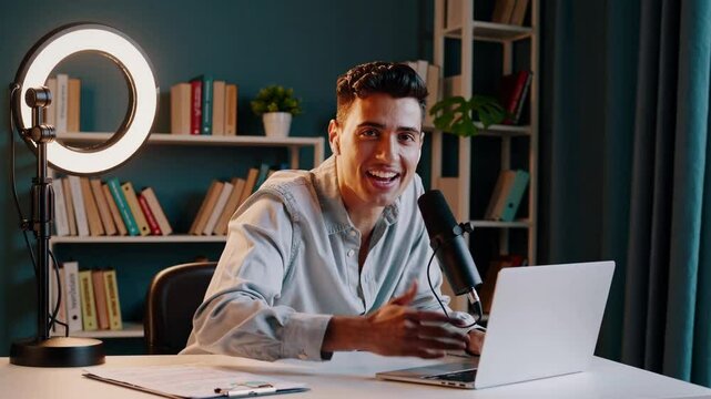 A cheerful man sits at a desk with a laptop and microphone, surrounded by a ring light and bookshelves, presenting as a video content creator in a cozy home studio