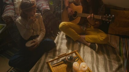 Full high angle shot of young Caucasian retro couple relaxing at home, man sitting on bed with crossed legs, playing guitar and singing, while woman is enjoying song, smiling and drinking tea - Powered by Adobe